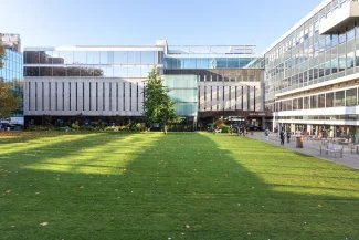 Photo of Queen's Lawn, Imperial South Kensington Campus, on a sunny day. The photo is mainly grass and the Library is in the background with a blue sky overhead.