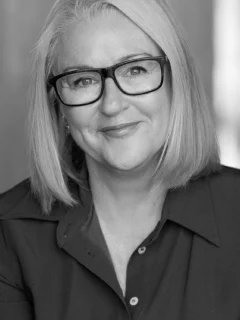 Black and white headshot of Jane, she has short blonde hair and is wearing a dark collared shirt and black rimmed glasses