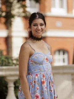 Nathalie is standing outside and wearing a blue, floral, sleeveless dress with a black necklace