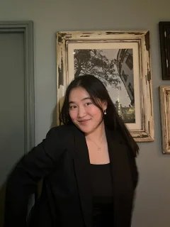 Monica is wearing a black top and jacket, and is standing in front of a wall with framed portrait photographs.